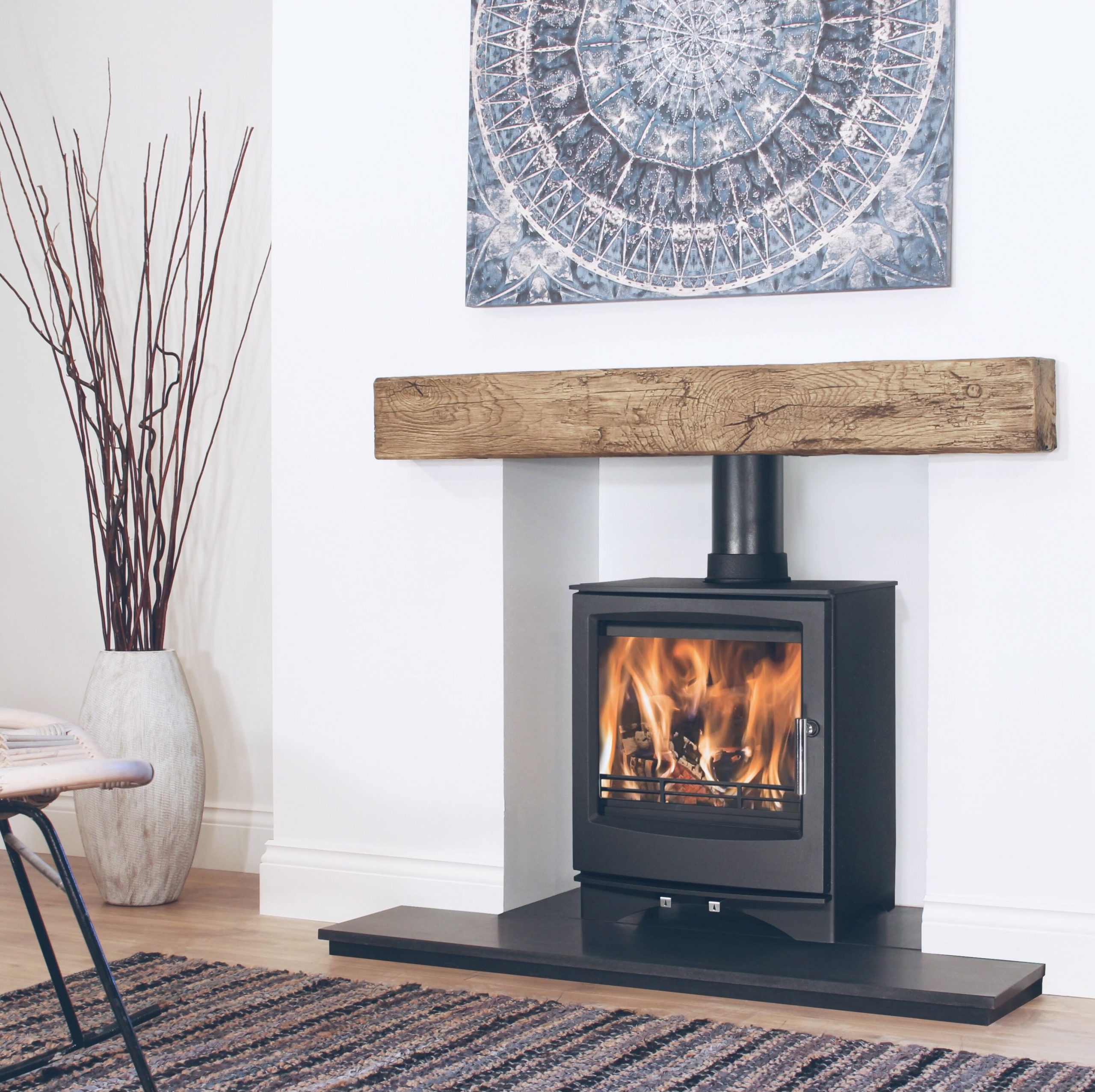 A lit wood burner in a modern room with a wooden mantlepiece, white walls and a mandala art work hanging on the chimneybreast.